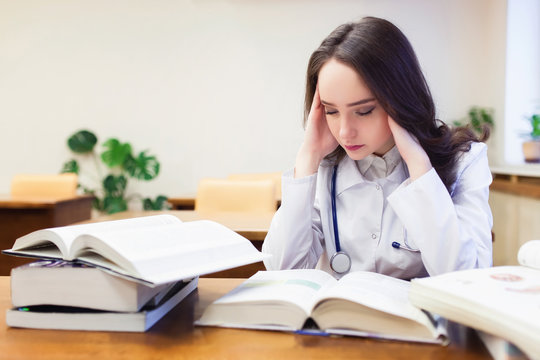 A female medical student with a headache is studying a new topic on textbooks.