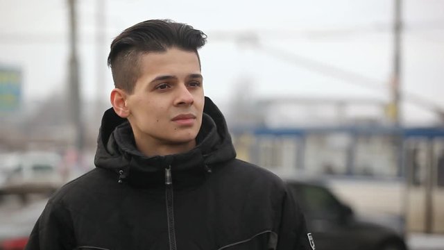 An impressive view of a young brunette man with a crew haircut  in a black anorak. He stands on a street and waits for his cutie girl in winter