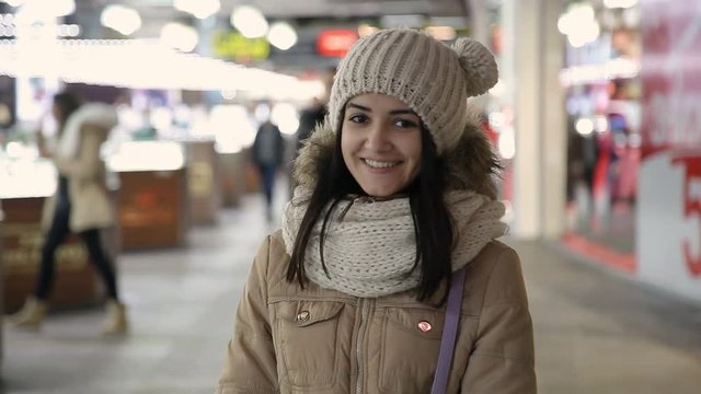 A wonderful view of a cheerful young woman in a creamy knitted hat, a fur anorak, smiling and standing near a shopping-case on a winter street.