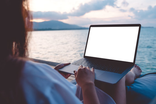 Mockup image of a woman using laptop with blank white desktop screen while sitting by the sea with blue sky background
