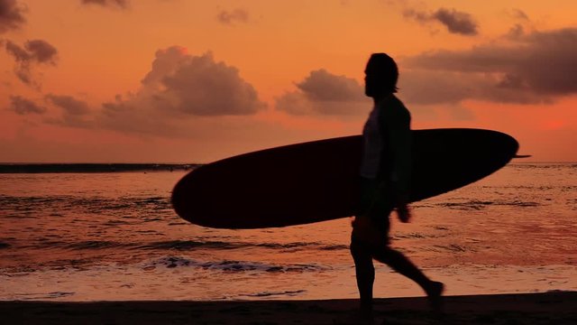 Side view silhouette of male surfer walking on beach carrying surfboard at sunset with orange sky on Bali island coast. Slow motion video