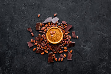 Chocolate and cacao concept. Cocoa powder in bowl near cocoa beans and broken chocolate on black background top view copy space