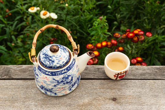 Chinese teapot and cup with green tea on a wooden table on a background of bright colorful flowers, top view. Pai, Thailand.