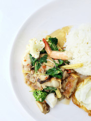 stir fried crispy pork, shrimp, mix seafood with thai holy basil, chilies, garlic, and soy sauce. in white dish with rice and thai fried egg style topping on white background.