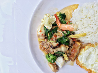 stir fried crispy pork, shrimp, mix seafood with thai holy basil, chilies, garlic, and soy sauce. in white dish with rice and thai fried egg style topping on white background.