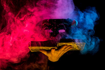 hand holding vintage book with mask in colorful smoke on black background