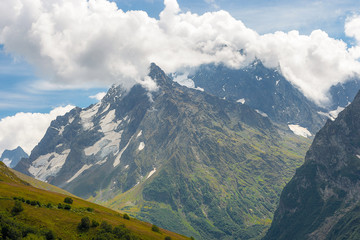 summer landscape of the Caucasus mountain peaks, green slopes in the grass with snow-capped peaks against the blue sky with clouds