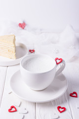 White cup with milk, homemade honey cake and red hearts on a white background. Free space