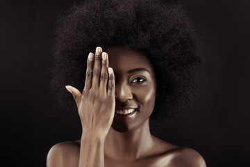 smiling african american woman covering eye with hand isolated on black