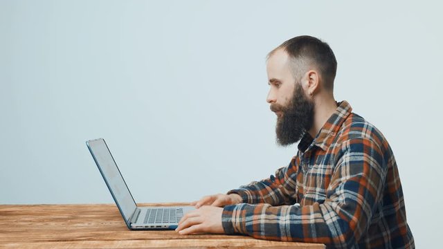 Bearded hipster man sitting at table working with laptop, turning to camera and smiling side view