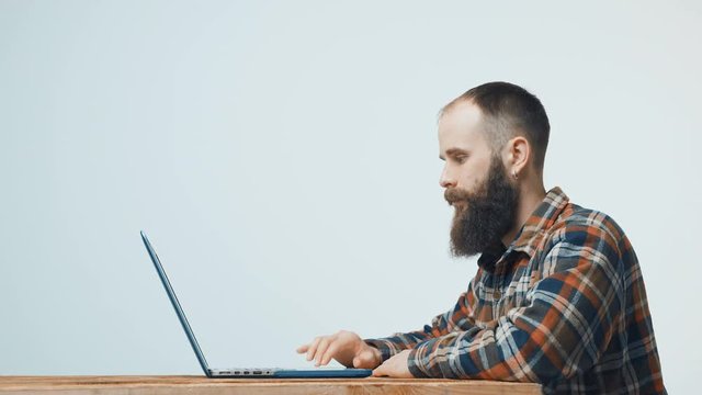 Bearded hipster man sitting at table working with laptop, turning to camera and gesturing thumb up, side view