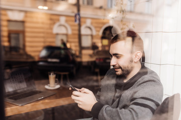 View through window with reflection street of confident man is typing text message on mobile phone in cafe