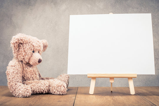 Sitting old Teddy Bear toy near portable desk easel for painting with canvas blank front concrete wall background. Retro old style filtered photo