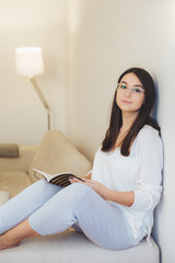 Obraz premium Photo of smart female student does test writing in notepad, prepares for examination while sits at comfortable sofa, wears glasses and casual clothes. Talented person works on creating design interior