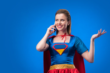 portrait of smiling woman in superhero costume talking on smartphone isolated on blue