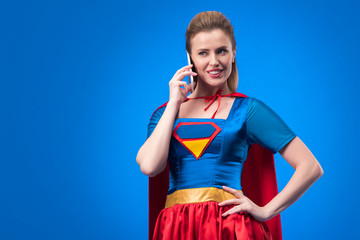 portrait of smiling woman in superhero costume talking on smartphone isolated on blue