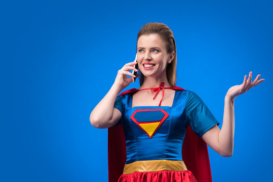 portrait of smiling woman in superhero costume talking on smartphone isolated on blue