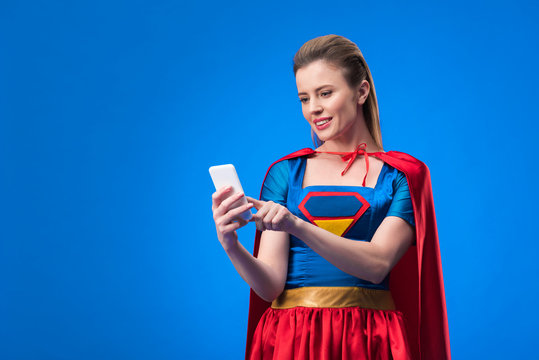 portrait of smiling woman in superhero costume using smartphone isolated on blue