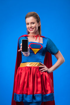 portrait of smiling woman in superhero costume showing smartphone with blank screen isolated on blue
