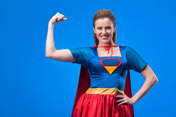 portrait of smiling woman in superhero costume showing muscles isolated on blue