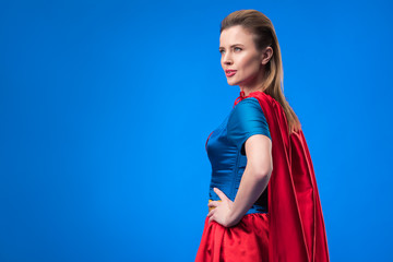 side view of beautiful woman in superhero costume standing akimbo isolated on blue