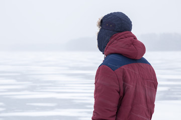 Fototapeta na wymiar Young man in snow blizzard standing alone on the lake ice and staring forward. Cold atmosphere in the afternoon. Foggy air. Winter concept.