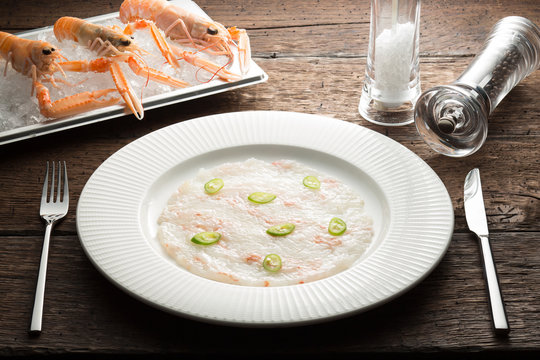 Scallops carpaccio served with langoustines on a white plate and wooden rustic brown  table with dinner settings. Close up 