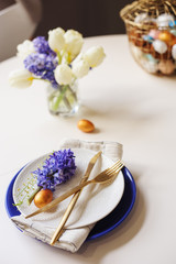 easter holiday dining table with golden metallic cutlery and eggs, hyacinth and tulip bouquet, blue and white plates. Celebration concept