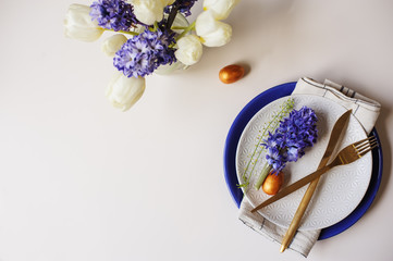 Easter holiday greeting card, dining table with plate, golden cutlery, painted eggs and hyacinth flower on white background with copy space. Flat lay, top view, trendy modern celebration style