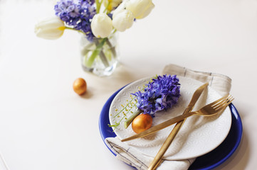 easter holiday dining table with golden metallic cutlery and eggs, hyacinth and tulip bouquet, blue and white plates. Celebration concept