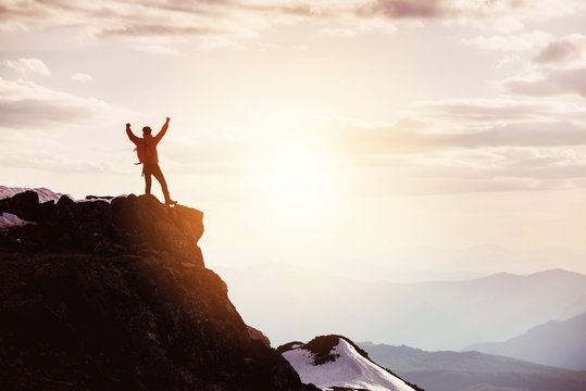 Man in winner pose at mountain top against mountains and sunset