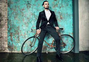 portrait of handsome fashion stylish hipster lumbersexual businessman model dressed in elegant black suit posing near sport bicycle and blue grange wall in studio. Metrosexual