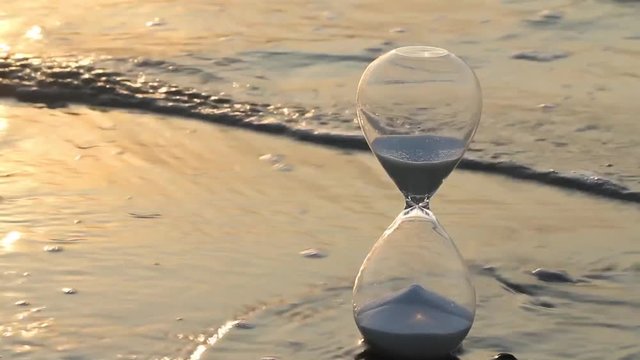 Hourglass on the beach touched by sea waves in bright and warm summer morning light showing starting time for a new day and running out in every moment