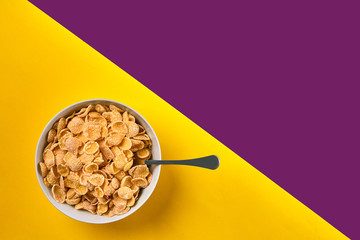 Bowl with corn flakes and spoon on purple and yellow background, top view