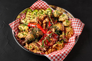 Assorted delicious grilled meat with vegetables and bbq sauce on an iron tray on black background close up. Big set of Hot Meat Dishes. Top view