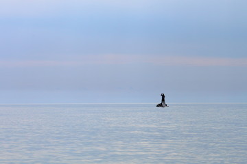 Silhouette of stand up paddle boarder paddling on a flat warm quiet sea. Water tourist with hermetic bags on SUP