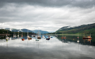 Obraz premium Boats moored on Loch Broom, Ullapool, Scotland. A view from Ullapool over the calm lake in the Scottish Highlands with overcast low cloud coverage.