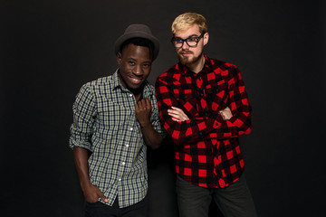 Studio shot of two stylish young men having fun. Handsome bearded hipster in a shirt in a cage standing next to his African-American friend in hat against a dark background.