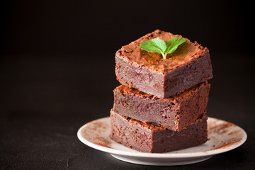 Chocolate brownie square pieces in stack on white plate with walnuts, decorated with mint leaves and cocoa on black background. Delicious dessert. Dark mood. Close up photography. Selective focus
