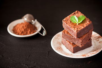 Chocolate brownie square pieces in stack on white plate with walnuts, decorated with mint leaves and cocoa on black background. Delicious dessert. Dark mood. Close up photography. Selective focus