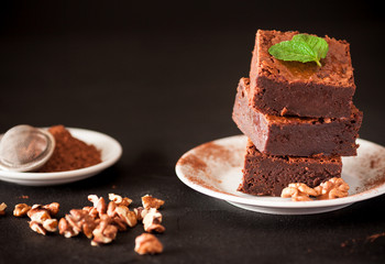 Chocolate brownie square pieces in stack on white plate with walnuts, decorated with mint leaves and cocoa on black background. Delicious dessert. Dark mood. Close up photography. Selective focus
