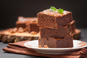 Chocolate brownie square pieces in stack on white plate with walnuts, decorated with mint leaves and cocoa on black background. Delicious dessert. Dark mood. Close up photography. Selective focus