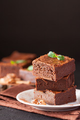 Chocolate brownie square pieces in stack on white plate with walnuts, decorated with mint leaves and cocoa on black background. Delicious dessert. Dark mood. Close up photography. Selective focus