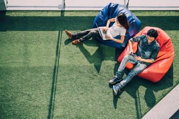 Young Asian couple using laptop notebook and digital tablet together in modern public park, sit on bean bag, top view with copy space on grass. Information technology gadget or casual business concept - Powered by Adobe