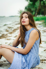  Beautiful young girl sitting on the seashore. Girl with long hair in a blue dress near the sea. tourist girl admires the sea view. Travel Concept. Time for rest and relaxation. 
