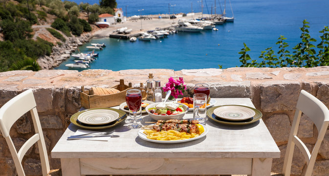 Fototapeta A table served for two with chicken souvlaki and french fries, greek salad, snacks and drinks on the summer terrace of the hotel room by the seascape, beautiful summer greek holidays concept.