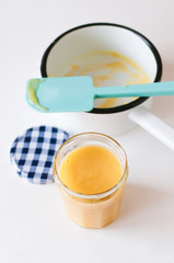 Close up of glass with homemade, traditional British lemon curd with sauce pot and turquoise silicone spatula on white background