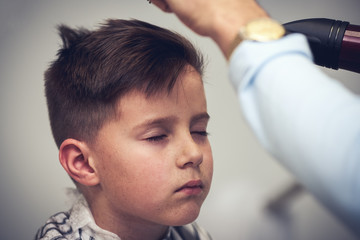Barber is drying a hair to Caucasian boy in barbershop.