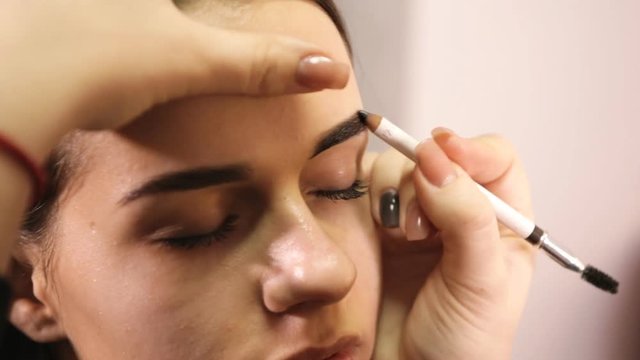 The hand of the makeup artist uses the pencil to draw eyebrows for the model