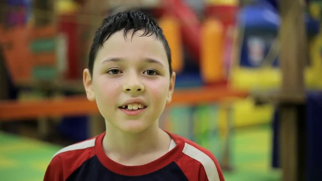 An optimistic view of a small sportive boy dressed in a red and black Tshirt. Though he is covered with sweat he smiles happily in a gym. 